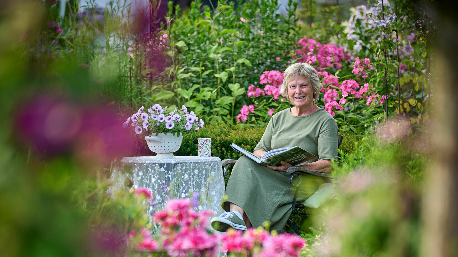 Garten, Blumen blühen in pink, Frau sitzt mit Buch an einem kleinen Tisch